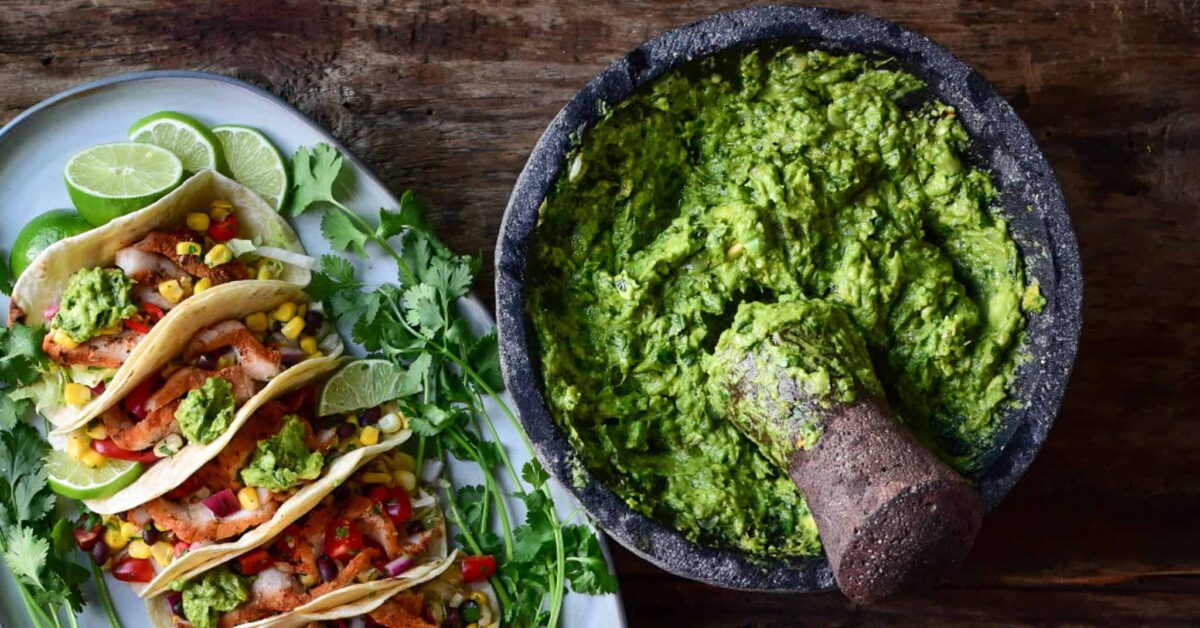 A large molcajete with freshly made grilled guacamole with a plate of tacos in the background.