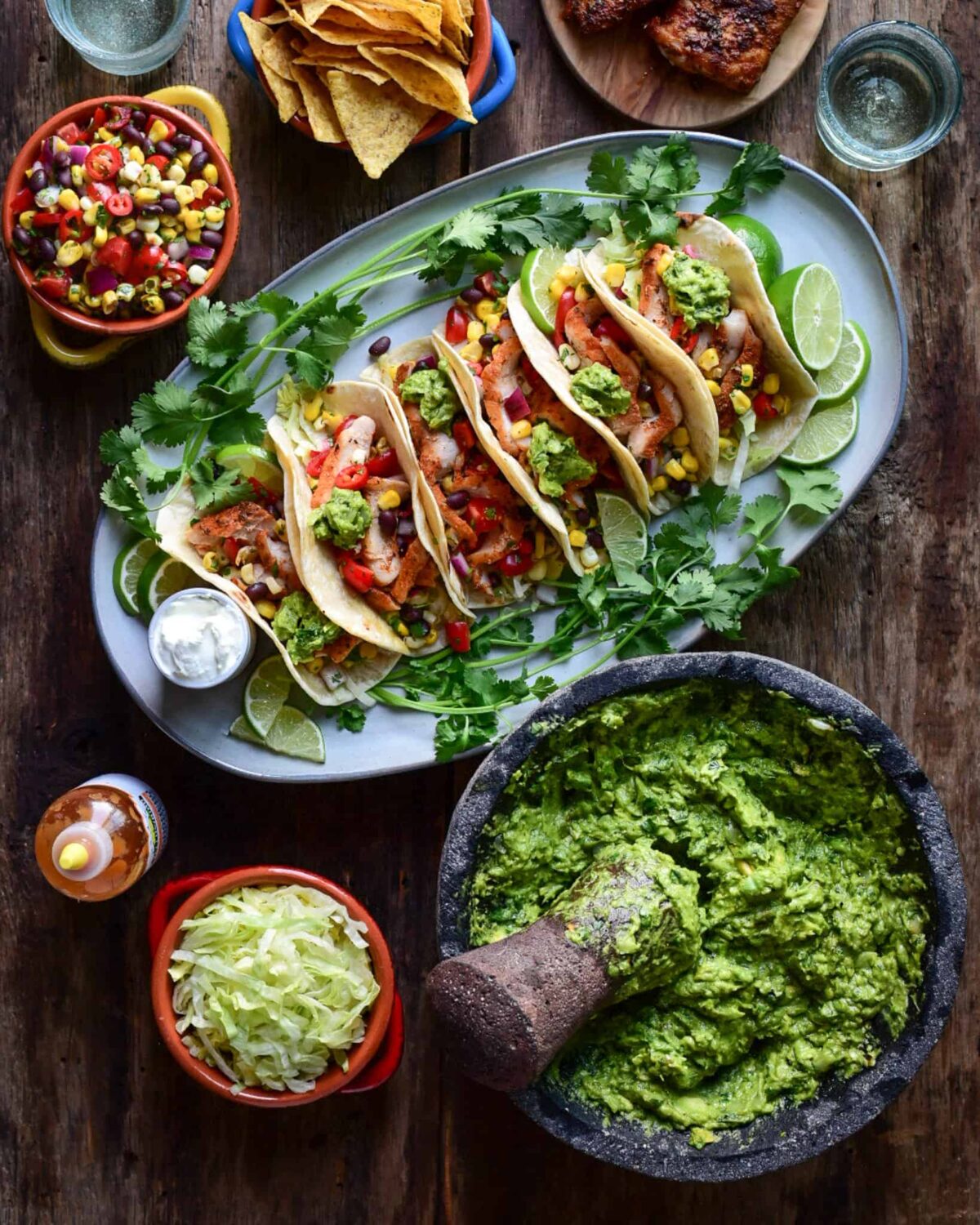 A platter of chicken tacos, a mortar and pestle with guacamole, served with bowls of various fixings.