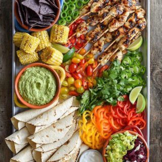 A sheet pan filled with grilled chicken skewers, tortillas, peppers, jalapeños, corn, cucumber and tortilla chips.