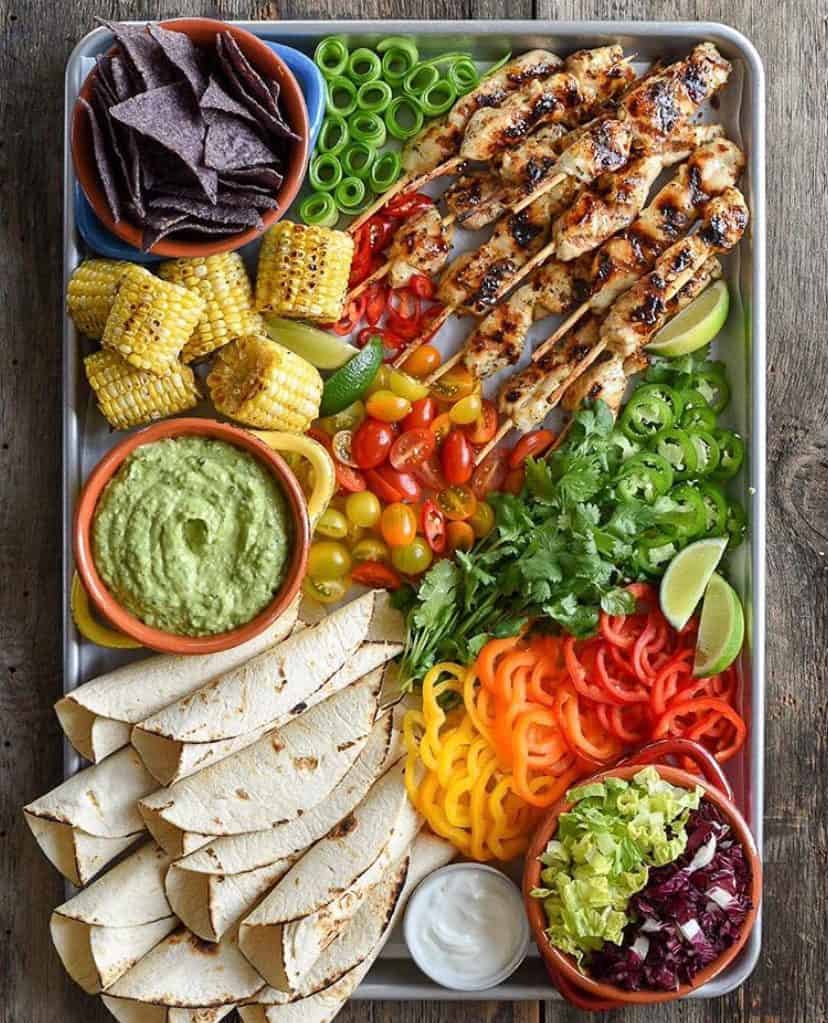A sheet pan filled with grilled chicken skewers, tortillas, peppers, jalapeños, corn, cucumber and tortilla chips.