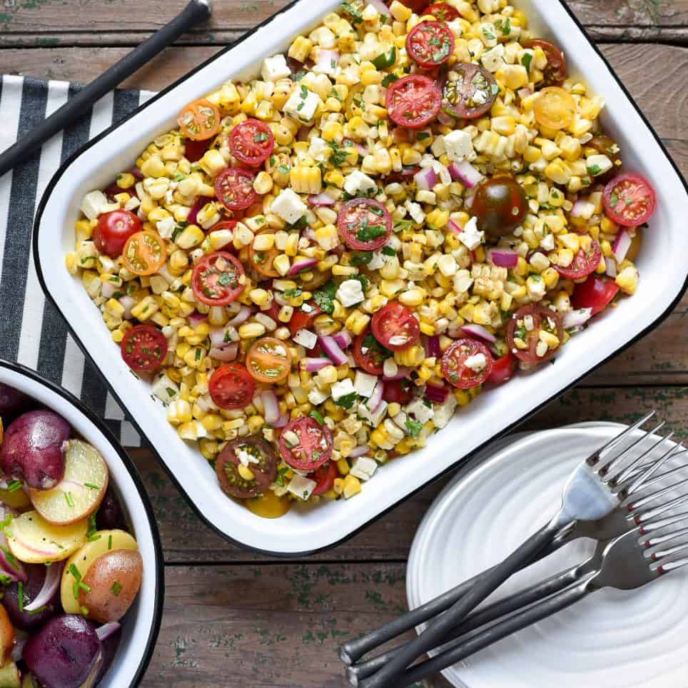 Rectangular white enamel tray with a grilled corn, tomato feta and onion salad. Forks and plates are ready for serving. 