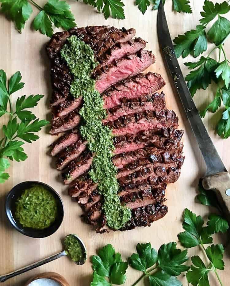 A sliced flank steak with a strip of chimichurri sauce down the top. Surrounded by parsley and a large knife. 