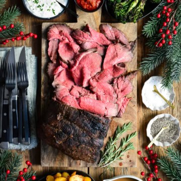 A large medium rare roast beef on a cutting board. Half of the roast is sliced into thin pieces. Served with gravy, potatoes, horseradish sauce and broccolini.