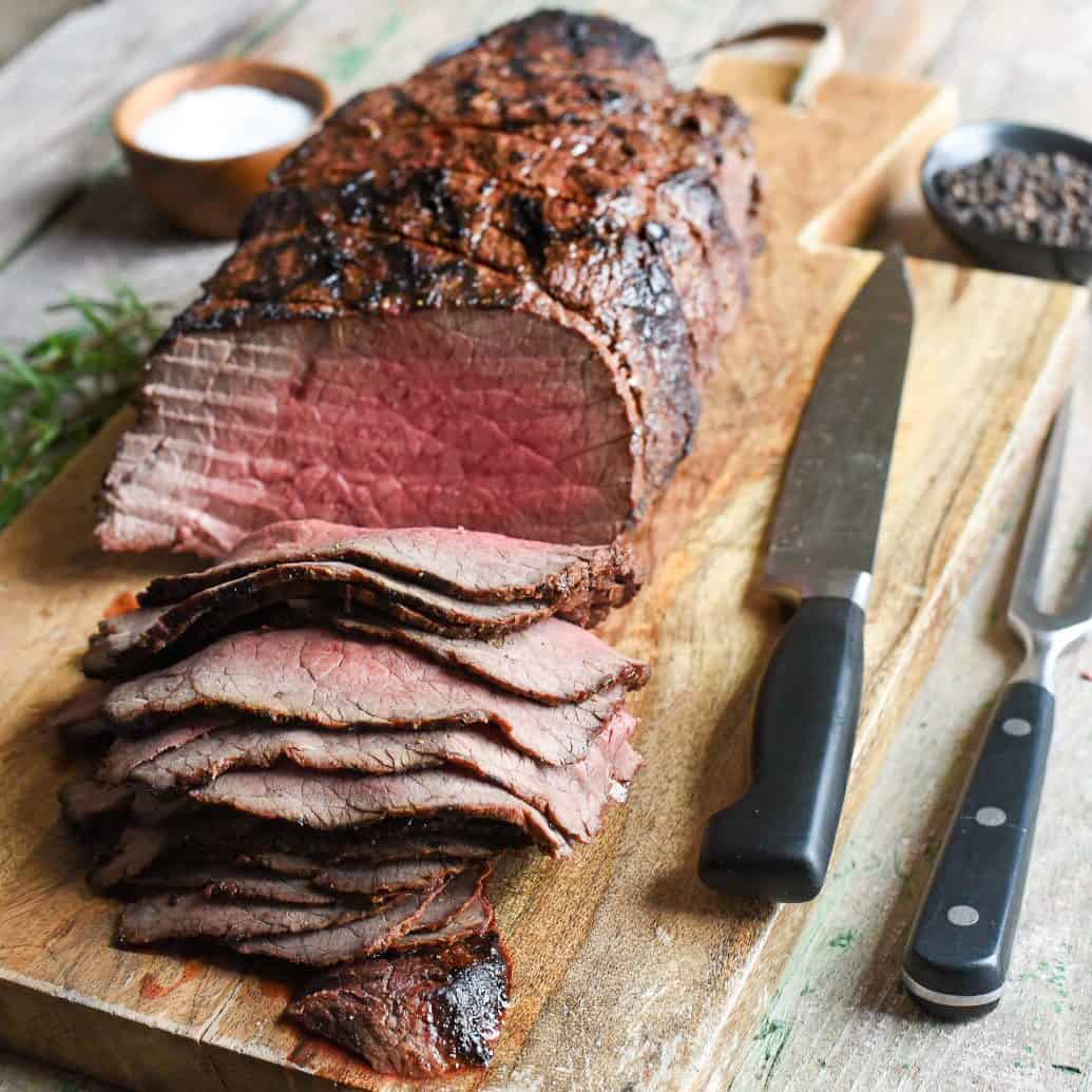 Grilled roast beef on a cutting board. Half of the beef is sliced thinly, a carving knife and fork are on the board.