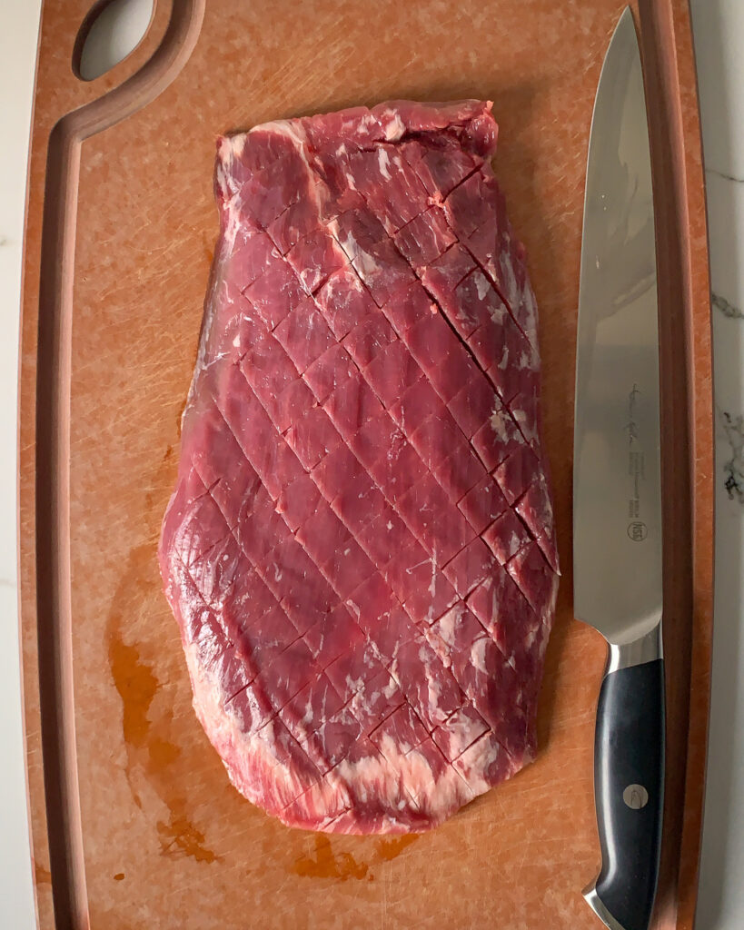 Raw flank steak, scored diagonally across the steak on a cutting board with a large Thomas Keller Cangshan  Cutlery knife.