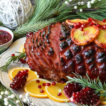 Side view shot of a glazed ham sliced garnished with pomegranates, rosemary and sliced oranges. Surrounded by Christmas decorations and a bowl of cranberry sauce.