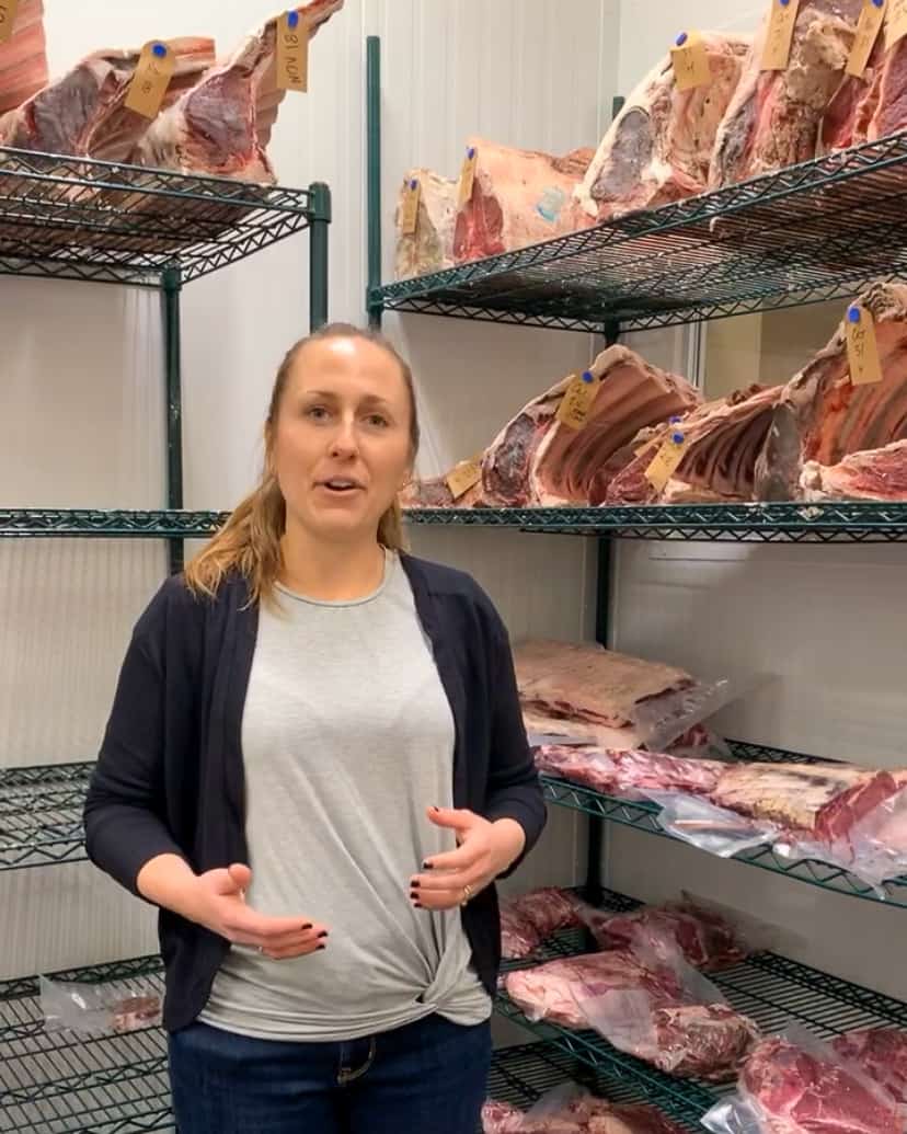 A woman in the butcher's fridge talking about the cuts of beef.