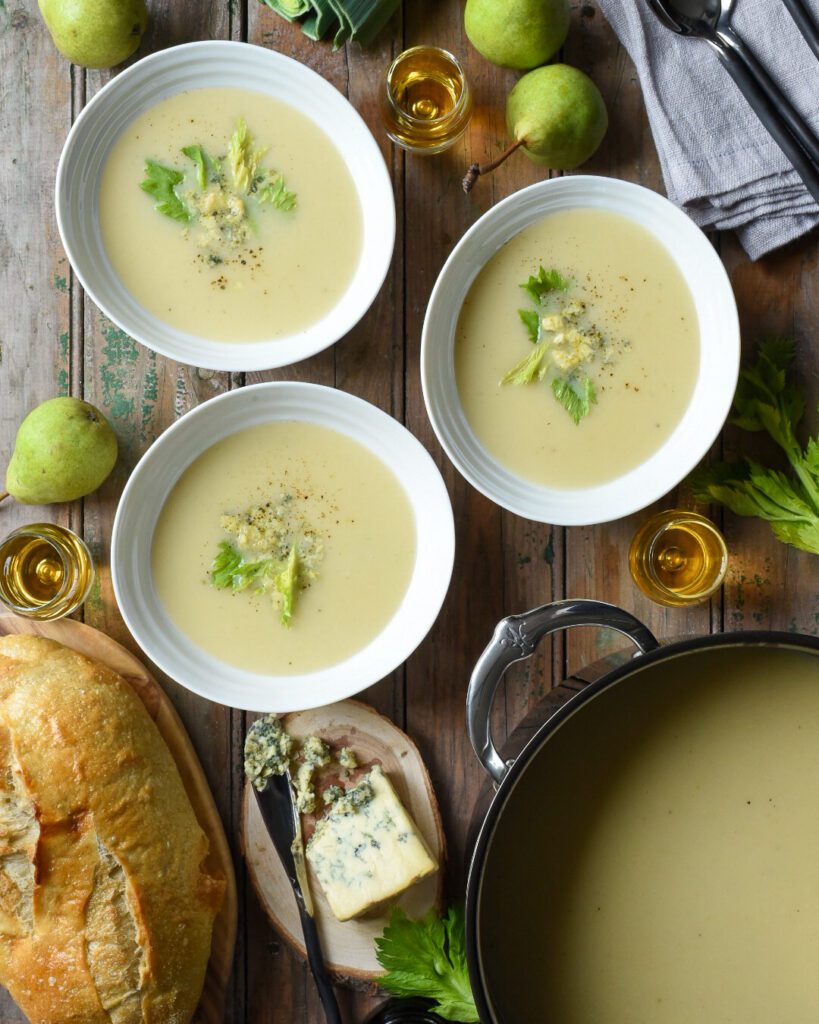 A large pot of  Pear, Port, Potato & Stilton soup with three bowls filled with garnishes. Three glasses of port, pears, cheese and bread are also on the table 