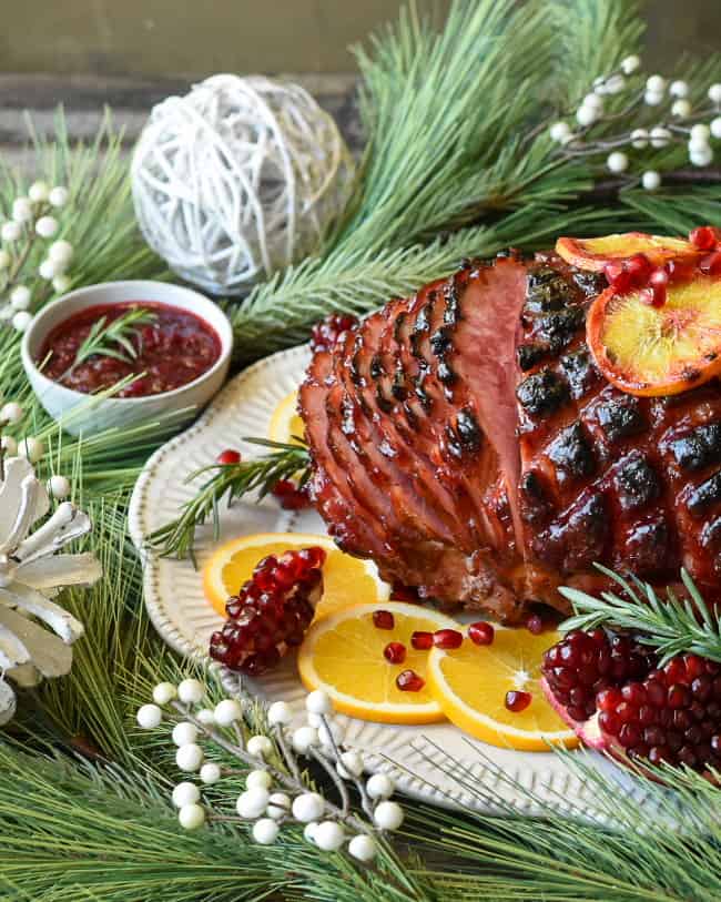 Side view shot of a glazed ham sliced garnished with pomegranates, rosemary and sliced oranges.  Surrounded by Christmas decorations and a bowl of cranberry sauce. 