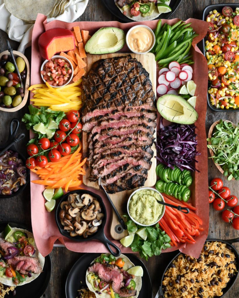 A grilled and half sliced flank steak is on a platter surrounded by a variety of colourful fresh chopped vegetables and taco toppings,