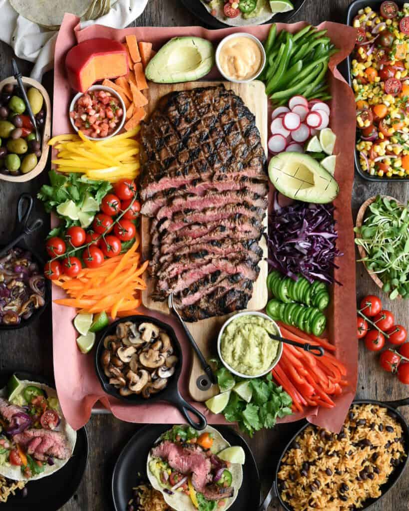 Top down view of a flank Steak sliced with taco ingredients surrounding. Ingredients include avocado, radishes, cabbage, jalapenos, sauted mushrooms in a mini cast iron skillet, guacamole, tomatoes on vine, mixed olives, cheese wheel with red wax, corn and tomato salad, rice and beans, and two tacos filled on black plates.