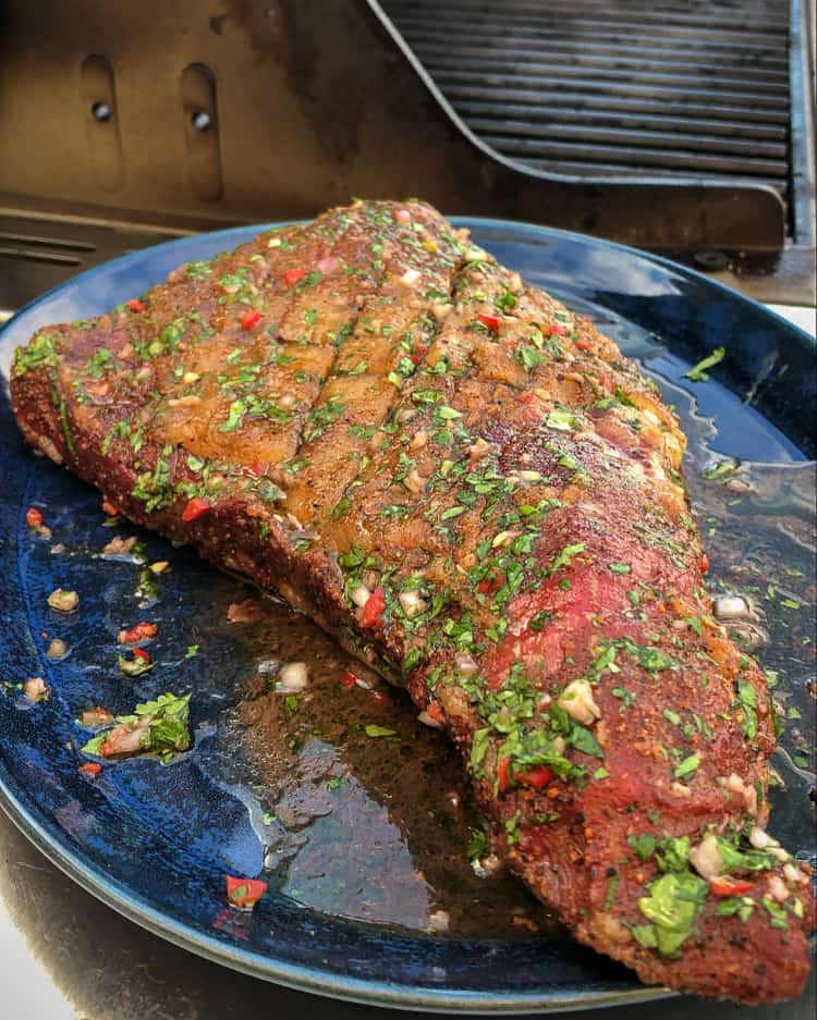 Tri-tip steak with marinade of chimichurri on a blue plate beside the barbeque about to get seared. 