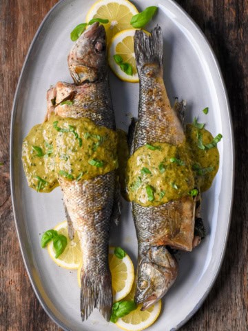 Two grilled branzinos on a plate with lemon slices and jalapeño-basil vinaigrette.