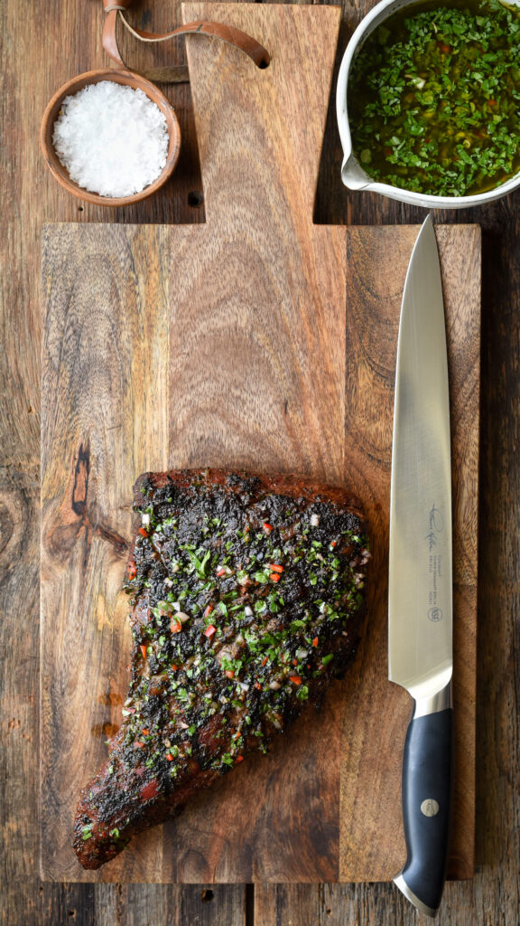 Tri-tip steak on a wooden board with a large knife. Bowl of chimichurri and a bowl of salt. 