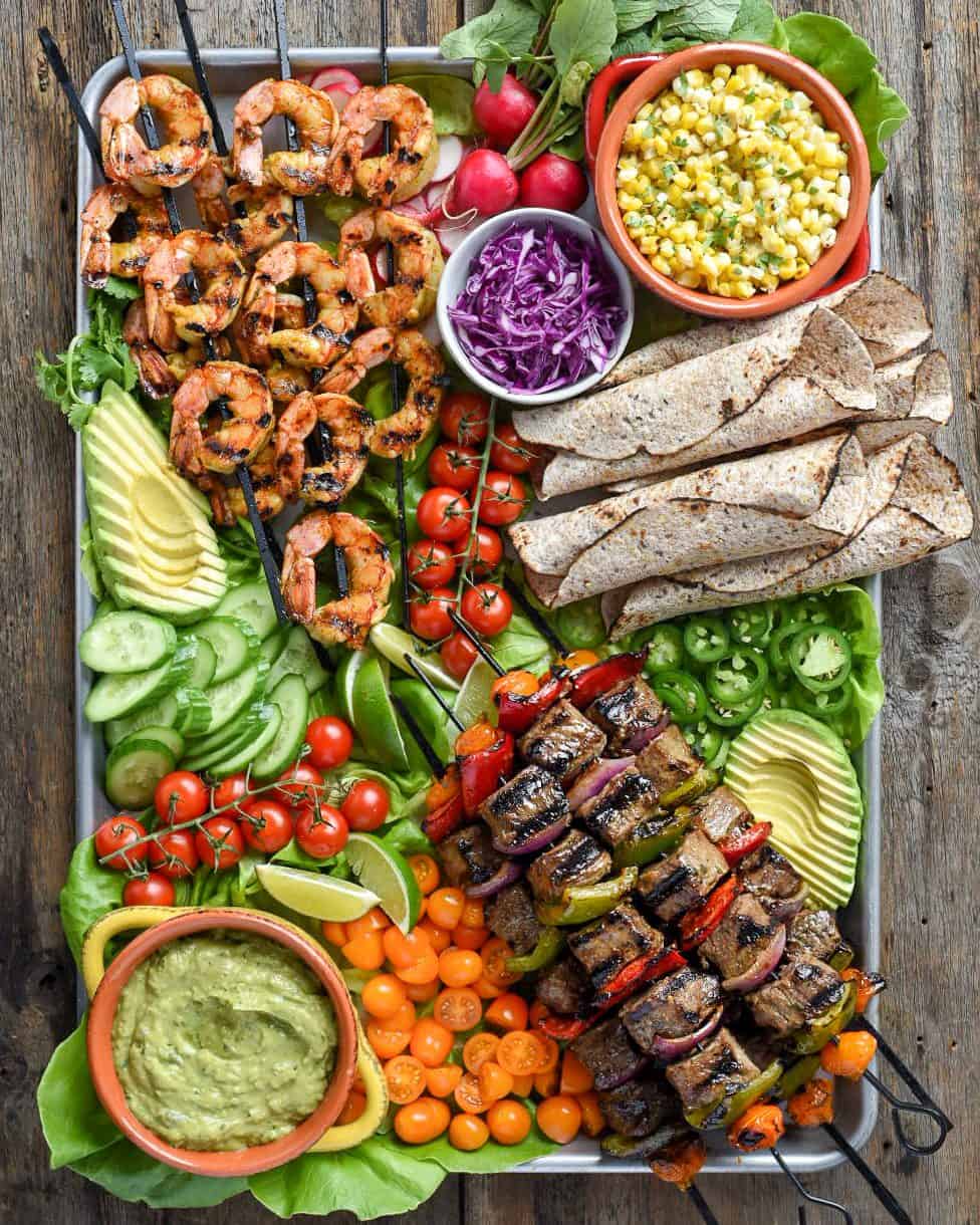 A large and colourful Surf & Turf platter of shrimp and beef skewers alongside grilled tortillas, spicy guacamole, tomatoes, avocados, sliced jalapenos, grilled corn and tomato salad, lime wedges and fresh chopped cilantro.