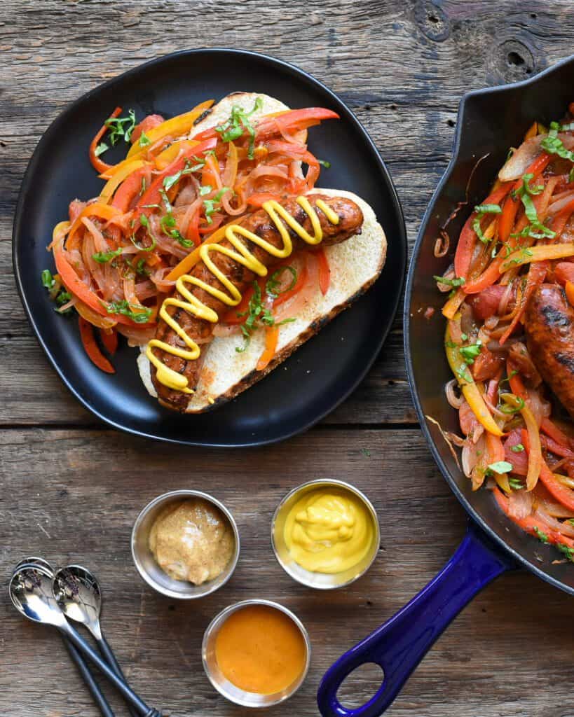 Ontario pork hot Italian sausages with peppers and onions in a blue cast iron skillet and on a bun on a black plate.  Three bowls of mustard.