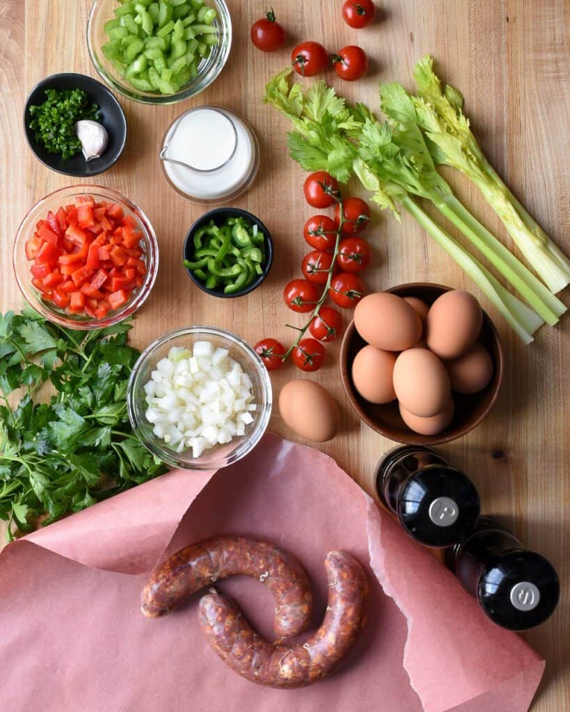 Ingredients for frittata laid out on a wooden board including, hot Italian sausage, eggs, tomatoes, peppers, milk, celery, salt and pepper.
