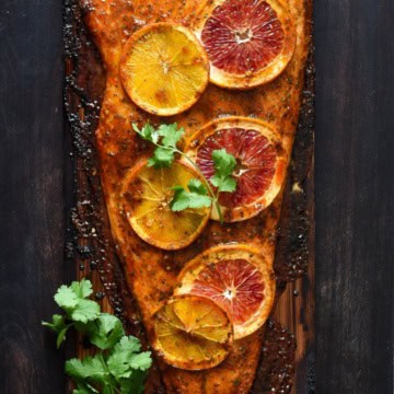 A cooked salmon fillet on a cedar plank with orange slices and cilantro.