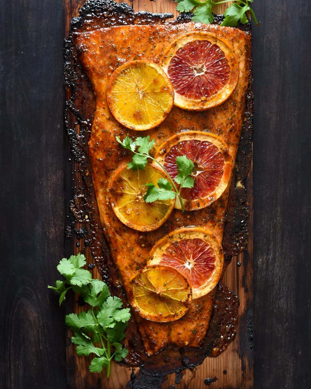 A cooked salmon fillet on a cedar plank with orange slices and cilantro.