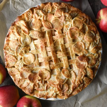 A baked apple pie, decorated with lattice and cut-out leaves on the crust. The pie is surrounded by several Ambrosia Apples.