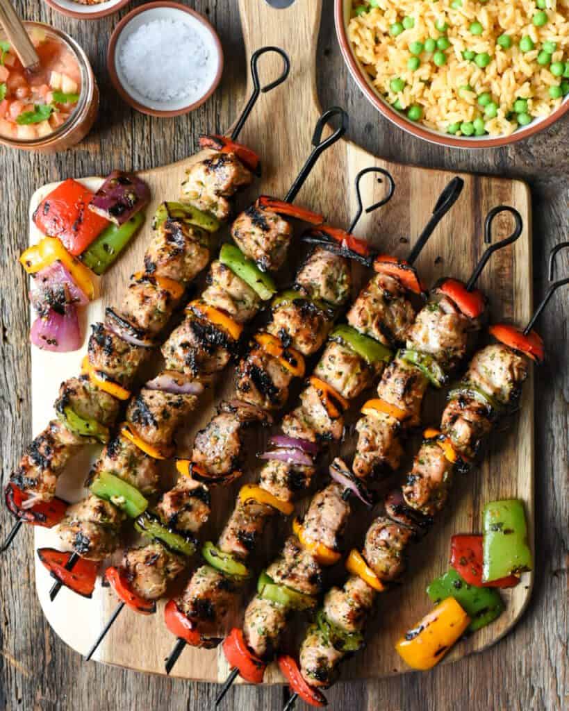 Six colourful, grilled pork tenderloin skewers with veggies on wooden board. Served with rice and peas and salsa.