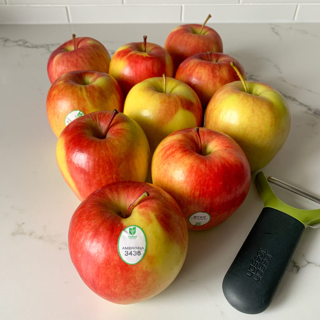 Ten Ambrosia apples with a peeler on the counter. Foodland Ontario tag is on the Ambrosia apple.