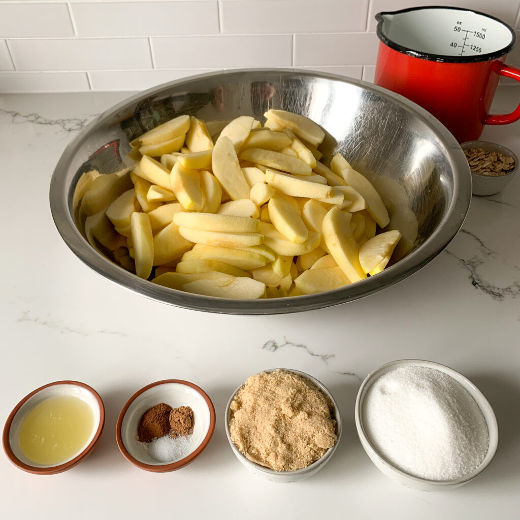 Large stainless steal bowl with sliced apples and four bowls of pie filling ingredients. 