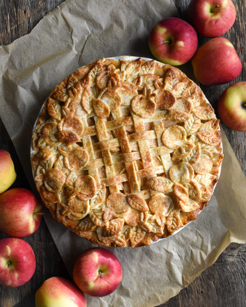 A baked pie with a lattice, apple and leaf cut out design. Apples also surround the pie. 