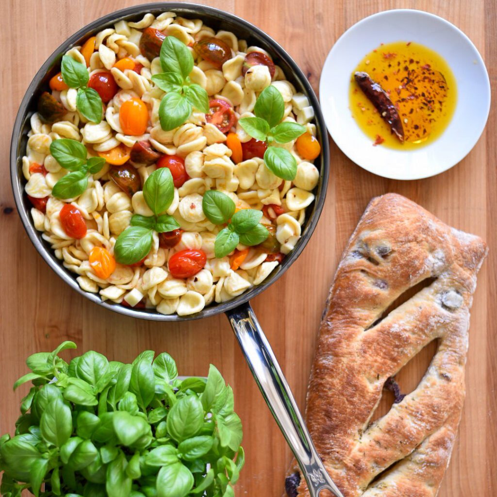 A large pan of Orecchiette with Heirloom Tomatoes, Mozzarella and basil. Surrounded by a dish of chili oil, a basil plant and an olive loaf. 