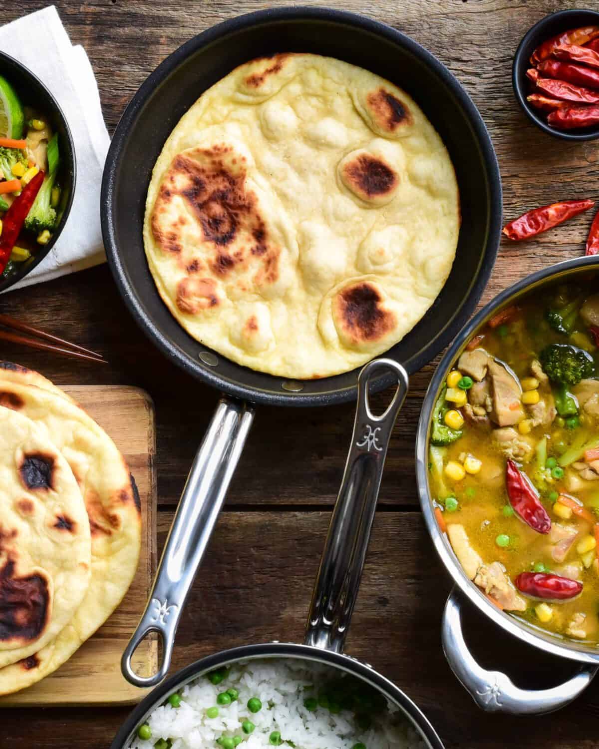 Top down image of a flatbread in a pan, a pot of Thai green curry, a pot of rice and peas and a stack of flatbread (naan).