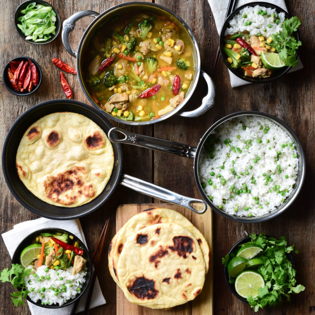A pot of Thai Green curry chicken, a pot of rice and peas, skillet flatbread and two bowls filled with the curry, rice and garnishes.