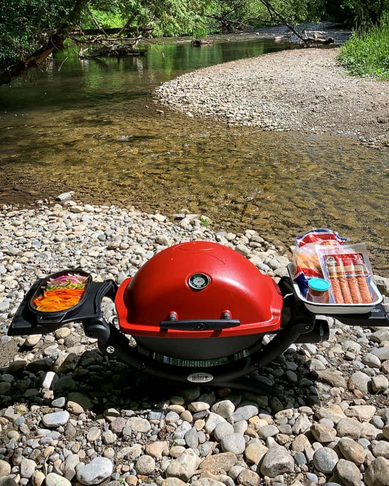 Red bbq with side arms out holding ingredients, by a river. 