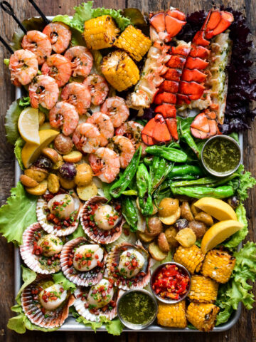 A large platter filled with grilled skewered shrimp, scallops in the shell, lobster tails, grilled corn on the cob and other fresh veggies.