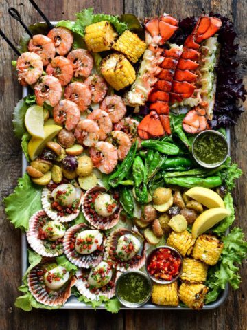 A large platter filled with grilled skewered shrimp, scallops in the shell, lobster tails, grilled corn on the cob and other fresh veggies.
