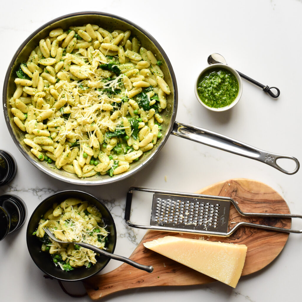 Top down view of a skillet of pasta surrounded with a bowl of pasta, pesto, cheese and a grater