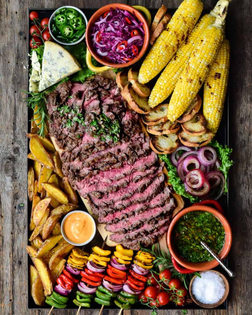 Platter filled with sliced flank steak beef, corn, potatoes, skewered veggies and chimichurri sauce.