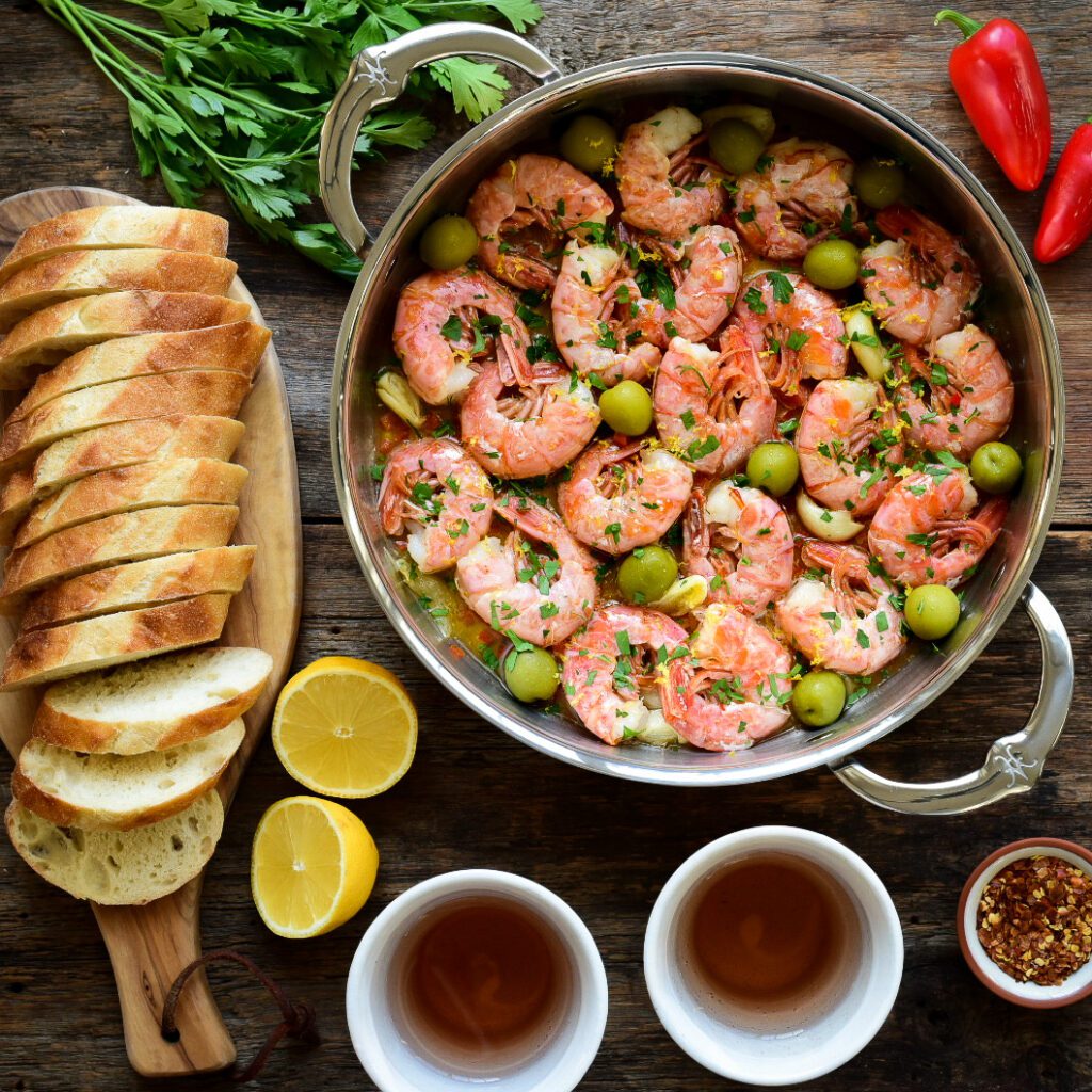 Top down image of pan with a garlic shrimp (Gambas al Ajillo). Surrounded by bread, wine, lemons and peppers. 