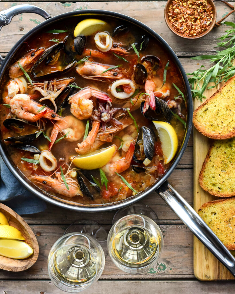 A top down image of a pan with Fisherman’s Stew with mussels.Toast, wine, lemons and crushed chilis surround this pan.