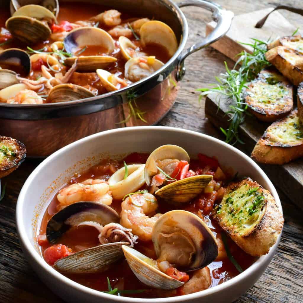 An images of a pot and soup bowl filled with Cioppino (Fisherman's Stew) with clams, shrimp and squid.  Garlic and herb toast in the background. 