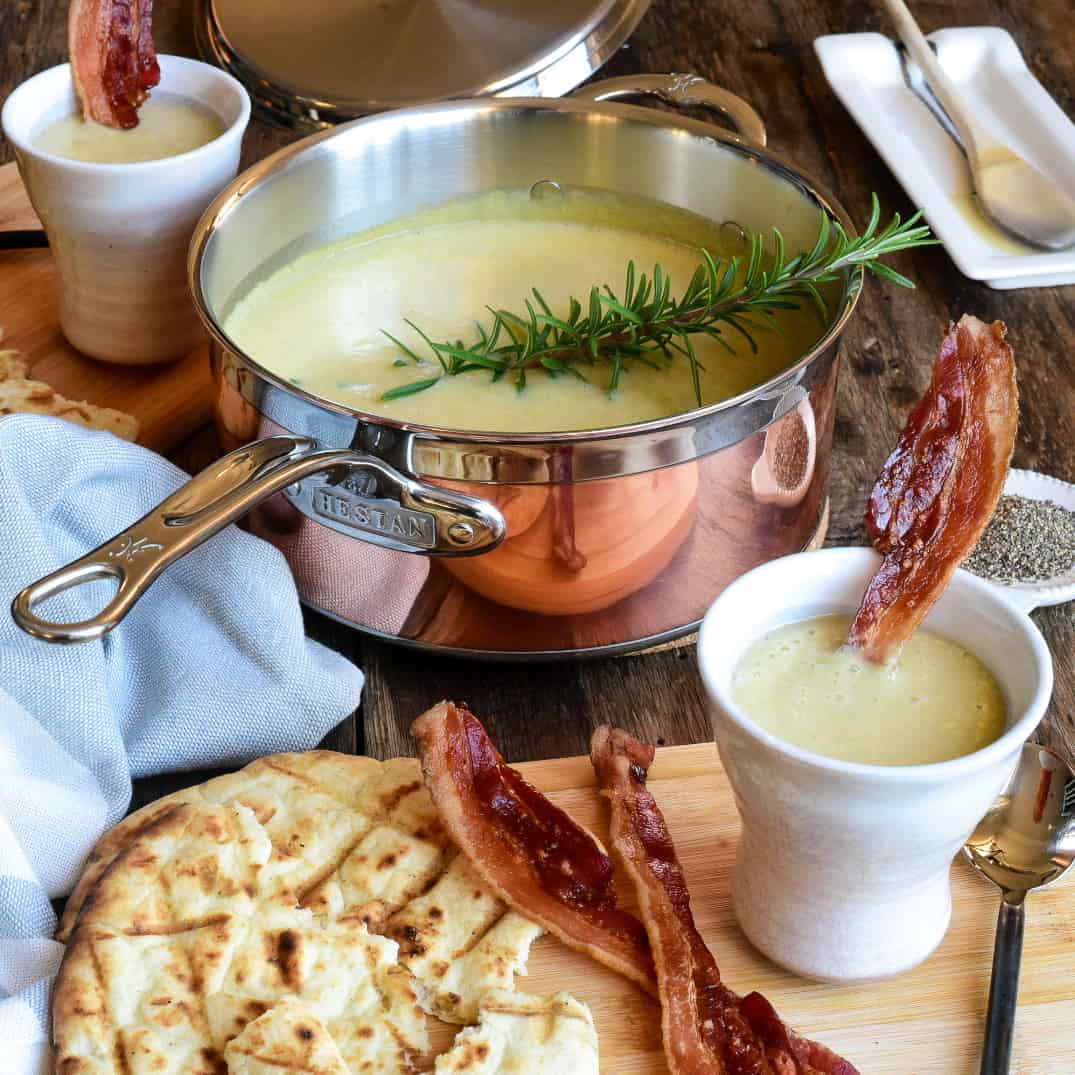 A copper pot with Leek Potato & Bacon Soup with a sprig of rosemary. Served in mugs with bacon and warm pita.