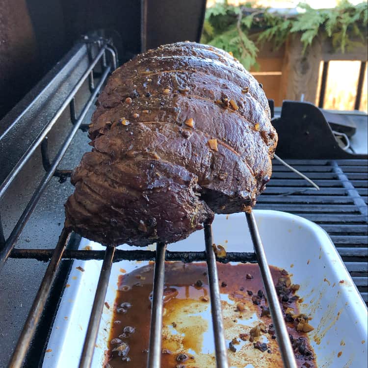 A finished beef roast on the top rack over the portion of the grill that is not on. A drip pan placed under the roast.