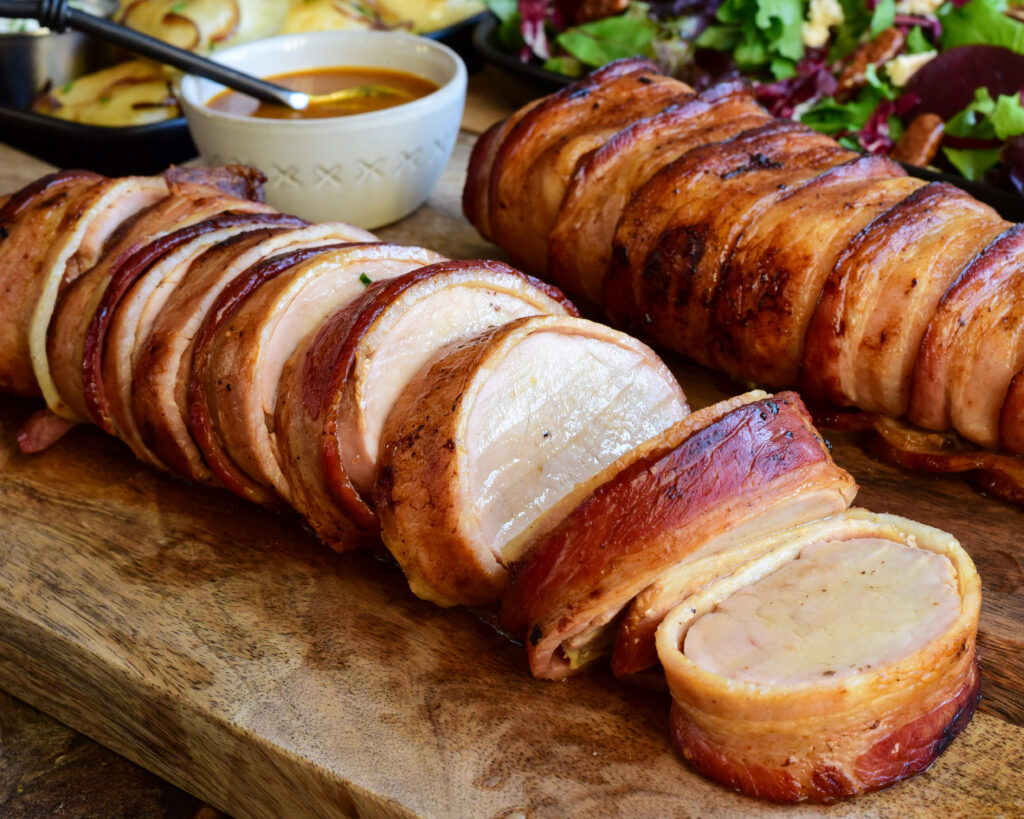 Two bacon wrapped pork tenderloins. One is sliced showing the juicy interior. 