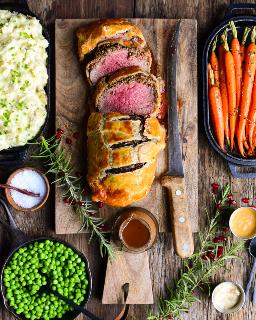 Top down image of a medium rare sliced Beef Wellington on a cutting board with a large carving knife and sauce. Carrots, peas and mashed potatoes surround the beef. 