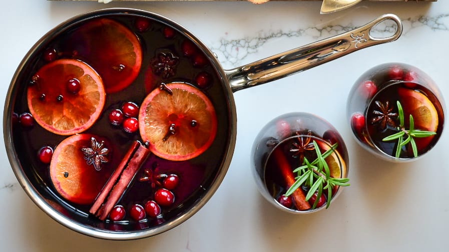 Top down image of a simmering pot of mulled wine and two glasses garnished with oranges, star anise, cinnamon and cranberries.
