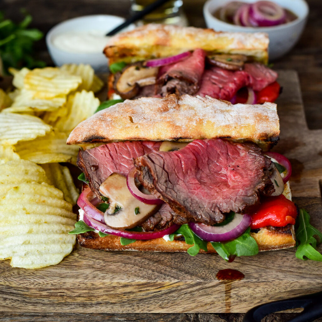 Roast beef Sichuan sandwich with red peppers, arugula, onions, sautéed mushrooms, horseradish sauce with a side of chips.