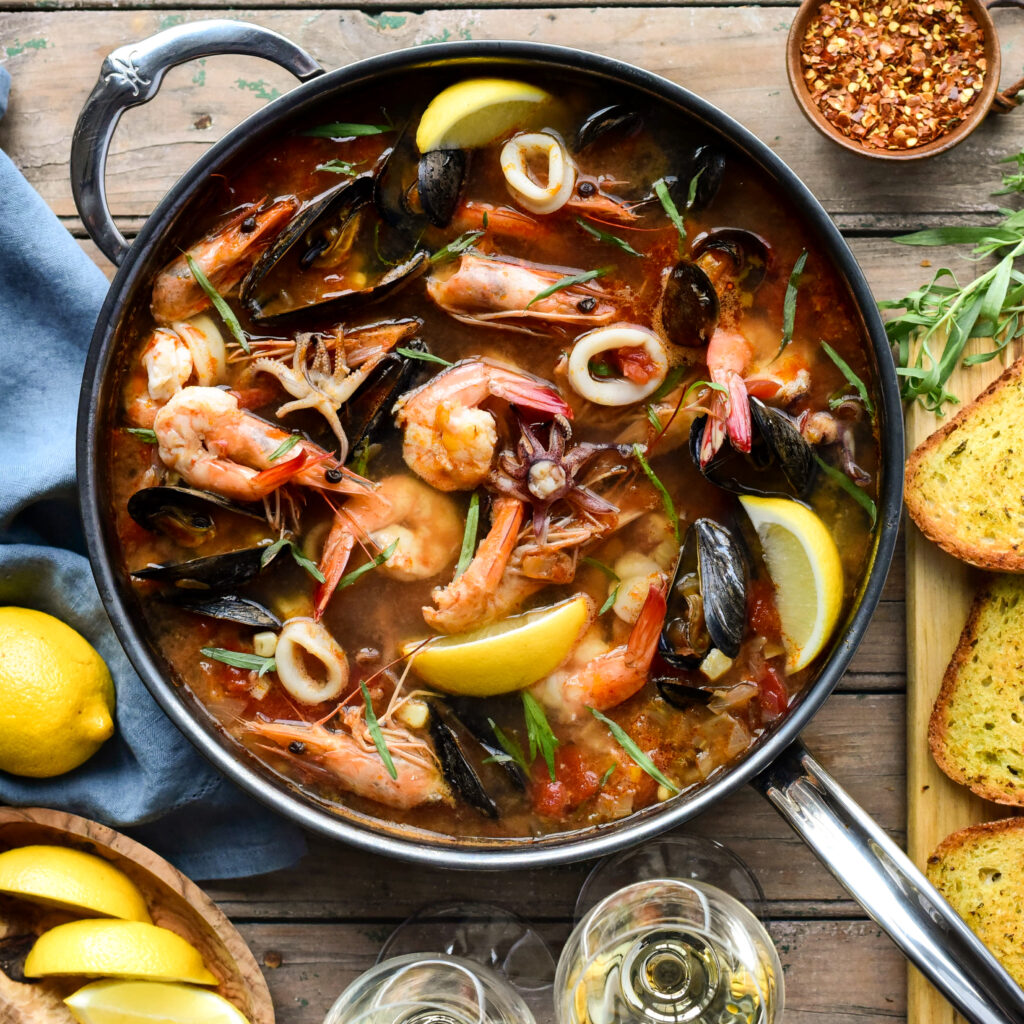 Top down image of a fish stew filled with mussels, shrimp, calamari, lemons and surrounded with wine glasses and garlic toast. 