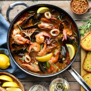 A top down image of a pan with a Cioppino (Fisherman’s Stew). Toast, wine, lemons and crushed chilis surround this pan.