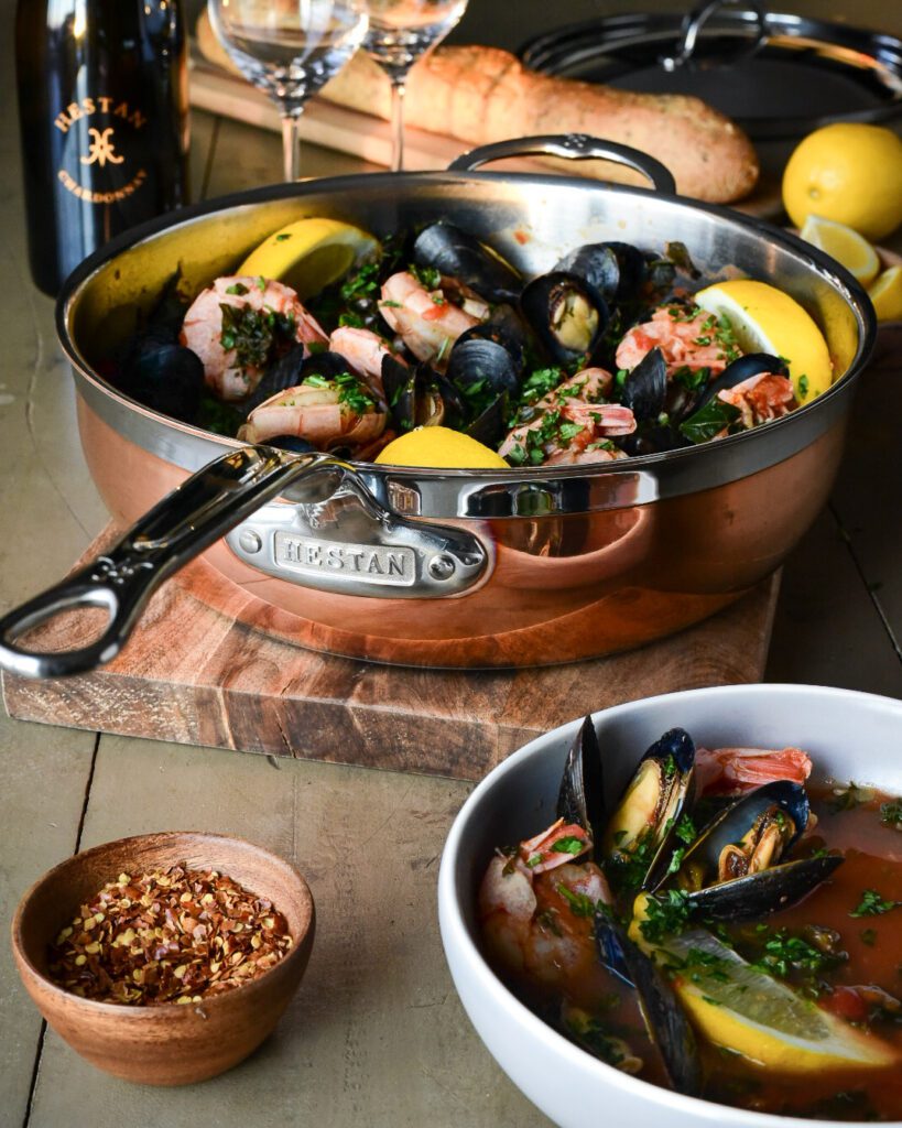A beautiful copper pot filled with mussels, shrimp, lemons and kale. This pot is surrounded by a bowl filled with this soup, a wooden bowl of chili peppers, wine, wine glasses, lemons and a loaf of bread.