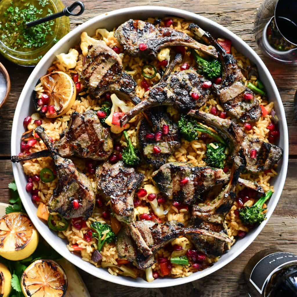 A top down image of a round platter of grilled lamb lollipops served over mixed vegetable rice and pomegranate arils. 