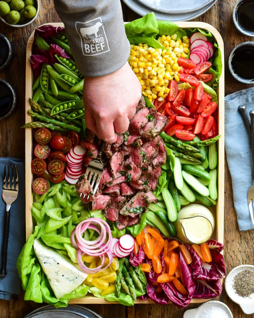 A bright and colourful grilled beef salad. Ribbons of medium rare beef are place in the middle of the tray and is surrounded by stack/piles of fresh vegetables, salad dressing and blue cheese. My arm is reaching in and I'm wearing a grey top with the Ontario Beef logo on the arm. 